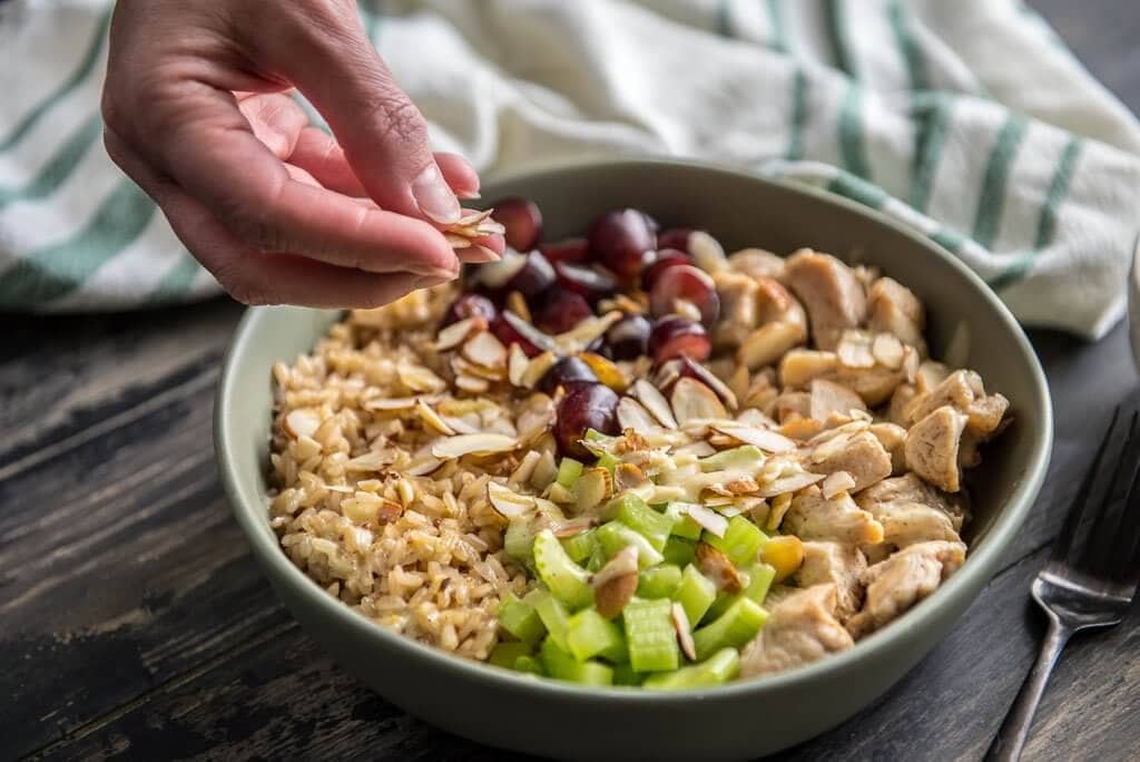 Bowl of healthy chicken salad brown rice bowl with grapes and celery and a woman placing almonds on top of the bowl.