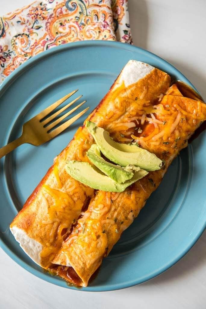 Serving of two pulled pork enchiladas on a blue plate with a napkin and a gold fork garnished with avocados
