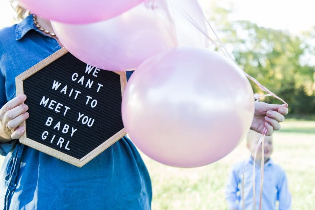 A woman holding pink balloons and a sign that says we can't wait to meet you baby girl