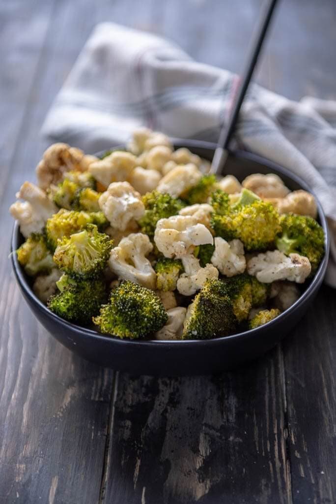 Roasted broccoli and cauliflower on a wooden table in a black bowl with a spoon and white napkin