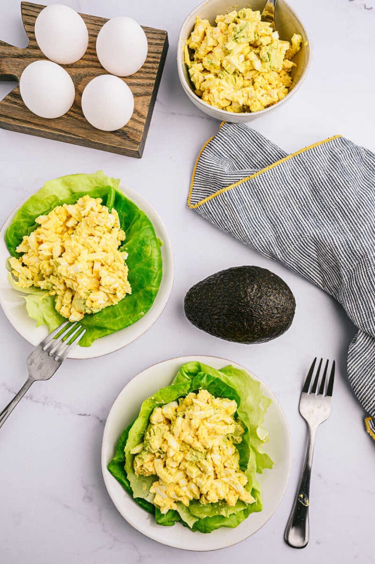 three bowls filled with avocado egg salad, one avocado a towel and eggs on a wooden egg holder. Shot from up above overlooking all of these items.