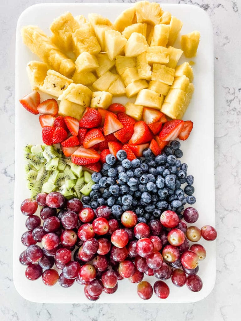 Grapes, pineapple, kiwi, strawberries and blueberries on a white cutting board on a marble countertop