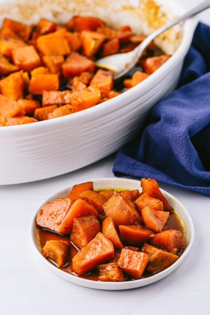 a small white dish of candied sweet potatoes casserole in front of the large baking dish filled with them and a blue napkin under the baking dish