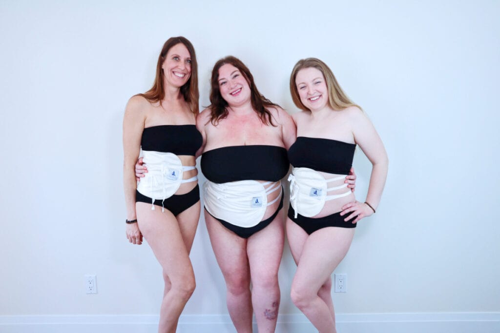 Three women standing together with black tube bras and black underwear and wearing castor oil packs over the right side of their stomachs