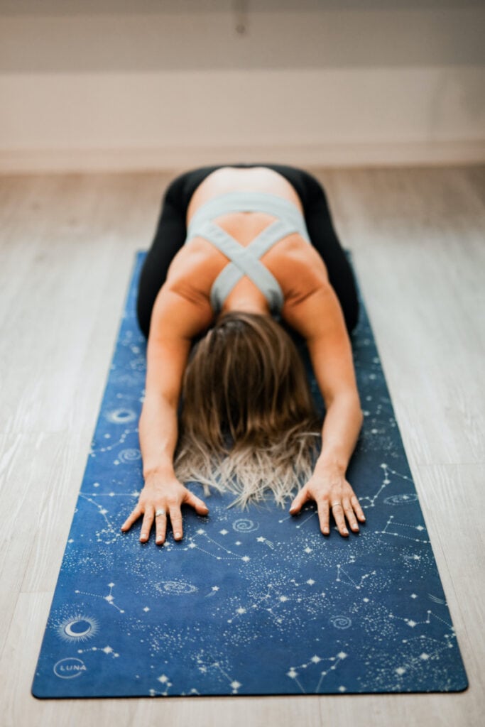 a woman in the yoga post childs pose on a blue yoga mat.  She is wearing black pants and a grey top and had blonde hair