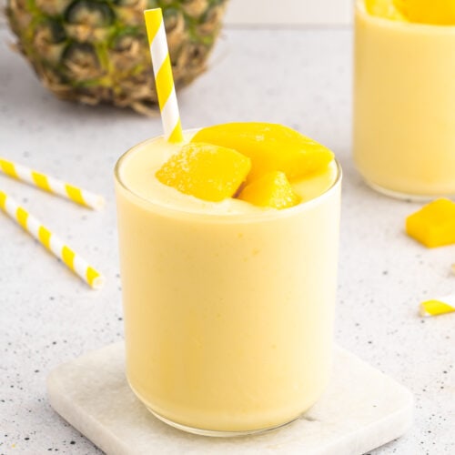 mango pineapple smoothie in a glass cup on a white coaster with a pineapple in the background and a straw in the cup