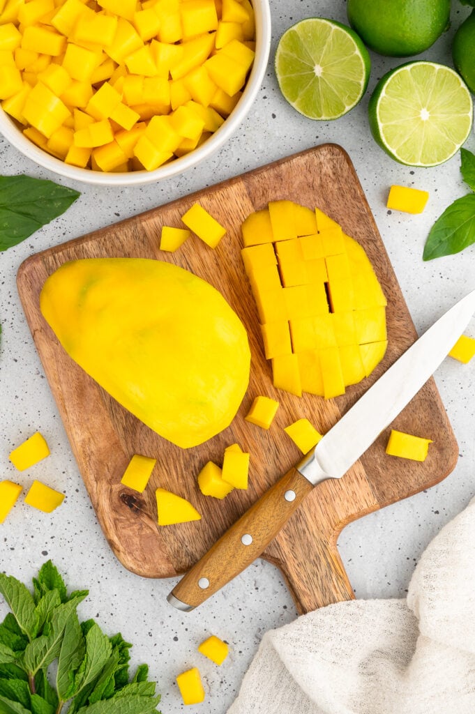 a wooden cutting board with one half of a full mango and the other half sliced with a paring knife