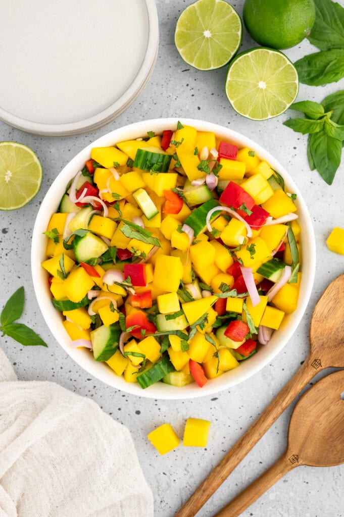 a bowl filled with cucumber mango salad with wooden spoons and limes