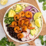 a bowl of shrimp, black beans, radishes, limes in a white bowl on a wooden charger on a white countertop
